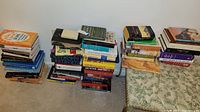 Multiple stacks of assorted books showing a variety of titles and subjects placed on a carpeted floor.