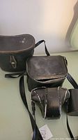 Photo shows two vintage camera cases with straps and an additional smaller camera or lens case in black leather, along with a black binocular case.