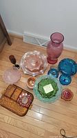 Photo showing assorted glass items on floor against wall, including tall pink swirl vase, iridescent bowls, green and blue bowls, amber trays.