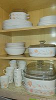 Shelves in a cabinet displaying CorningWare dishes with peach floral pattern. Includes stacked plates, bowls, mugs, and casserole dishes with glass lids.