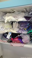 Photo of shelf with assorted folded fabrics in various colors and textures including white, purple, green, reds, some wrapped in plastic.