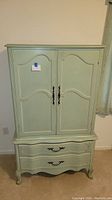 Full front view of the green painted wood armoire showing two doors and two drawers at the bottom with metal handles.