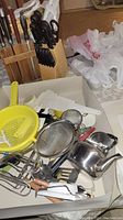 Overview of the lot showing a knife block with knives, kitchen scissors, a yellow plastic colander, metal strainers, whisks, spatula, and other small utensils in a bin.