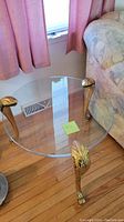 Clear circular glass top supported by three gold lion paw feet legs, photographed next to a couch and pink curtains on a wooden floor.