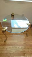 Round glass top end table with brass legs featuring shell-shaped decorative feet on hardwood floor.