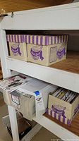 Photo showing two Crafts World cardboard boxes with purple stripes stacked on white shelf, containing sewing patterns.