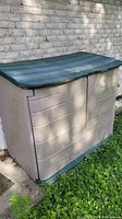 Photo of large plastic outdoor storage shed with beige body and green lid, placed against brick wall on concrete pad with grass nearby.