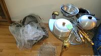 Main view of used pots, pans, metal cooking strainers, mesh racks, and cooking utensils with wooden handles arranged on floor and small table