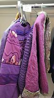 Three handmade jackets hanging on a rack varying shades of purple with textured and striped patterns.