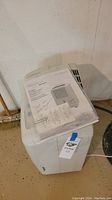 White Soleus portable dehumidifier with instruction manual on top, placed against a wall on a beige floor.