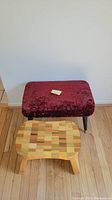 Two step stools placed against a white wall and wooden floor. One is a wooden step stool with a mosaic pattern top and four legs. The other is an upholstered footstool with dark red velvet-like fabric and black legs.