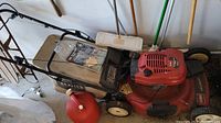 Full view of Toro red lawn mower with black handle, fabric grass catch bag, and white wheels, placed on a concrete floor near garden tools and a chair. Includes a red gas can and a cleaning brush near the mower.