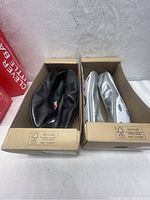 Two pairs of Puma ballet flats, one black and one silver, displayed inside their recycled cardboard boxes on white surface.