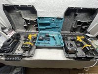 Three cordless drills in open carrying cases on a table, including two yellow and black DeWalt drills and one teal Makita drill.