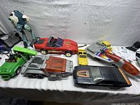Wide photo showing all toys on a table including red convertible, silver six-wheeled car, green truck with robot figure, black car, yellow Camaro, yellow face car, and retro rocket
