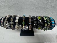 Front view of all 13 bracelets on black display stand, showing variety in colors and designs including black, green, blue, and clear bead styles with charms