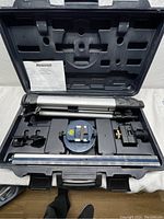 Photo showing the opened carrying case containing a silver adjustable tripod, blue laser leveling device with bubble level, mounting accessories, and a manual.