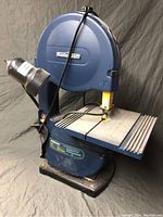Front angled side view showing the Mastercraft band saw, blue body, ribbed metal work surface, lamp attachment, and power cord.