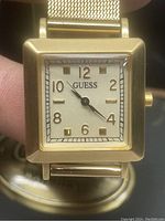 Front view of GUESS wrist watch showing square gold tone case, white dial with gold numbers and hands.