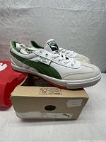 Side view of white and green PUMA TT Super CC sneakers on top of the shoebox with PUMA packaging in background.