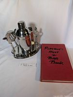 View of entire bar set next to the vintage Playboy's Host & Bar Book, showing all components including the cocktail shaker, bar tools on the stand, and the book closed on white background.