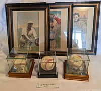 Lot showing three signed baseballs in glass display cases in front of four vintage framed baseball player pictures.