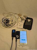 Photo showing Samsung Galaxy S6 with 'Welcome!' screen, two Coby MP3 players, sealed Google headphone adapter box, charging cables, wall adapter, and phone case.