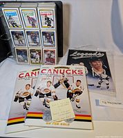 Photo of a black binder with hockey cards in plastic sleeves, two Vancouver Canucks coin albums and one Legends sports memorabilia magazine with uncut sheet in plastic wrap on a table.