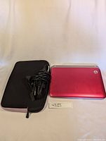 Closed red HP mini laptop placed next to a black soft laptop case and power adapter with cords coiled, on a white surface.