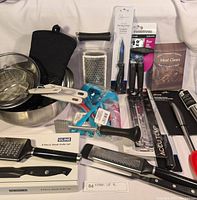 Full view of kitchen lot items laid out on a white surface showing metal mixing bowls, oven mitt, graters, knives, peelers, meat claws, meat thermometer, and other kitchen tools