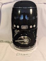 Front view of the black SMEG 50s style drip coffee maker showing the brand logo, digital screen, and control buttons.