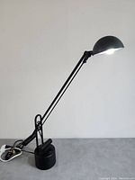 Black adjustable cantilever table lamp with dome-shaped head illuminated, showing side profile and power cord.