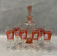 Seven-piece set of peach-colored glassware including a decanter with stopper and six stemmed cordial glasses decorated with white grapevine designs and gold accents.