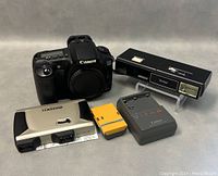 Canon EOS 20D camera body alongside vintage Vitara pocket camera, Vivitar flash, Canon charger, yellow accessory and stereo viewer laid out on neutral background.