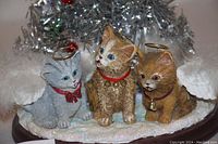 Close-up showing the detailed figurines of three angelic cats with halos and wings sitting around the base of a silver tinsel Christmas tree decorated with red and green baubles.