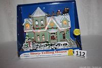 Full boxed view of Christmas story-telling house in green with snowy roof, Santa and animals figurines, yellow and red lights along roof.