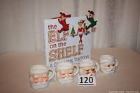 Photo showing four Santa mugs, two rubber elves perched on the book, and the book upright behind mugs.