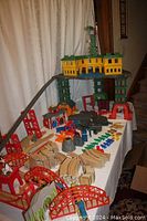 Wide view of the table with various wooden and plastic train pieces including the large yellow roundhouse with green roof and crane, multiple bridges and track pieces spread out.