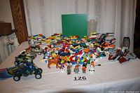 Wide view showing large pile of assorted Lego bricks, base plate, built walls, vehicles, and minifigures on a white surface against curtain background.