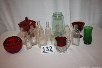 Lot of antique medicine bottles, colored glassware including red candy dish, ruby creamer, green vase, and mason jar on a white cloth background.
