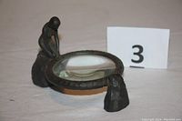 Angled front view showing entire magnifier resting on table with patinated metal frame and kneeling figure