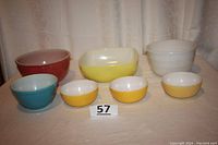 Seven vintage bowls and a yellow Pyrex square baker arranged side by side on a table to show colors and sizes.