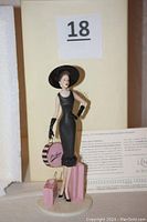 Front view of the Lenox porcelain figurine showing a fashionable lady in a black dress and hat, holding a pink round shopping box and two pink shopping bags on base.
