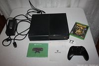 Front view of Xbox One console with controller, power supply, game case, and manuals laid out on white surface.