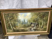 Framed acrylic painting of a forest scene with trees, a small cabin, and a stream, photographed front-facing with white backdrop.
