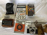 Photo of camera kit, showing vintage camera in brown leather case with strap, 35mm photography handbook, Tiffen filter set, Prisma lens cover and Vivitar filter box, with small plastic bags at top left