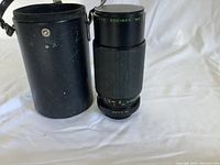 Makinon Auto zoom lens with an 80-200mm zoom range standing upright next to its black leather or faux leather carrying case with snap button and strap.