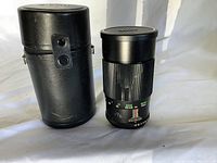 Photo showing black Auto Vivitar telephoto 200 mm camera lens with marked focal lengths and aperture settings, next to a black cylindrical lens case with button snap closure.