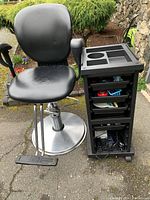 Black cushioned hairdressing chair and black mobile storage cabinet side by side outdoors on pavement