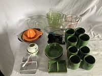 Box of kitchen utensils and dinnerware including measuring cups and spoons, lemon juicer, ceramic cups, bowls, fluted glass dishes, and drink coasters.